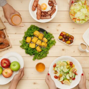 A table with a variety of foods including bread, donuts, apples, a salad, grilled corn, tomatoes, fried eggs with bacon, sausages, and two glasses of juice. Several hands are reaching for different dishes.