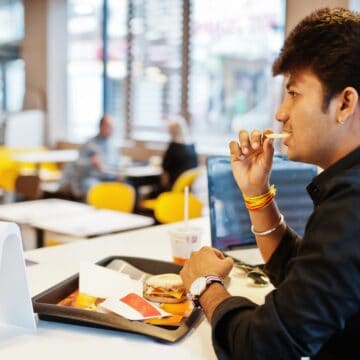A person sits at a table with a tray of fast food, including a burger and fries, in a restaurant. They are eating a fry and have a drink nearby. The table has a sign displaying the number 29. There are other patrons in the background.