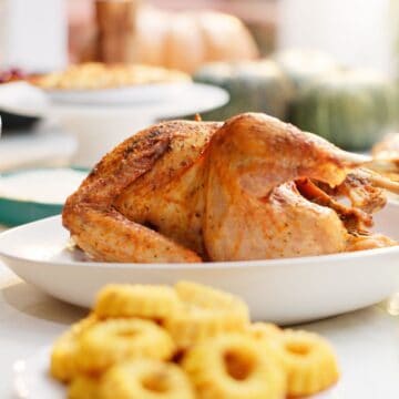 A roasted turkey takes center stage on a white platter, flanked by festive side dishes like curly fries and greens. In the background, blurred pumpkins hint at holiday foods. The feast promises indulgence while being mindful of options high in sodium for those enjoying this celebratory meal.