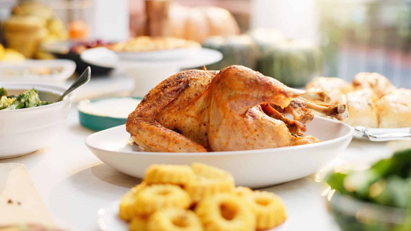 A roasted turkey takes center stage on a white platter, flanked by festive side dishes like curly fries and greens. In the background, blurred pumpkins hint at holiday foods. The feast promises indulgence while being mindful of options high in sodium for those enjoying this celebratory meal.