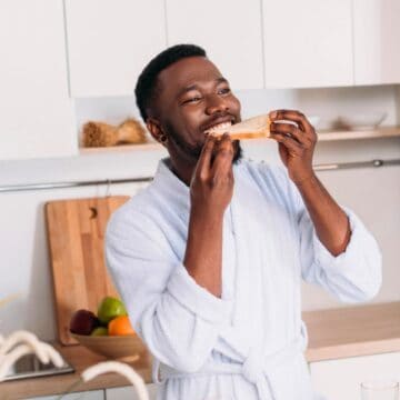 A man in a white bathrobe stands in a kitchen smiling while eating a slice of toast. In the background, there is a countertop with a cutting board, a bowl of fruit, and white cabinets.