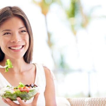 A woman is sitting outdoors, smiling while holding a plastic bowl filled with a fresh salad. She is holding a fork with salad greens. There is a blurred background of greenery and sunlight.