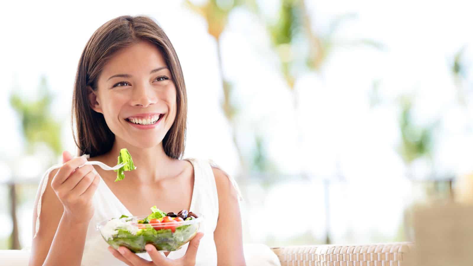 A woman is sitting outdoors, smiling while holding a plastic bowl filled with a fresh salad. She is holding a fork with salad greens. There is a blurred background of greenery and sunlight.