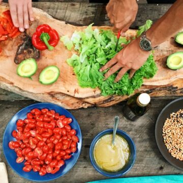 Two people preparing food on a rustic wooden surface. Ingredients include chopped red peppers, leafy greens, halved avocados, sliced cucumbers, cherry tomatoes, pine nuts in a pan, and a bowl of dressing. A knife and a bottle are nearby.