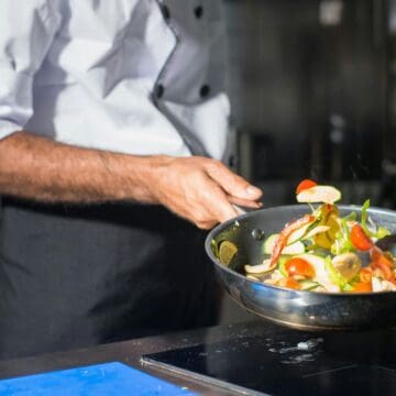 A chef in a white jacket and black apron is holding a frying pan, expertly tossing assorted vegetables like cherry tomatoes and peppers, showcasing creative ways to incorporate more vegetables into meals. The kitchen background features stovetops and a tiled wall.