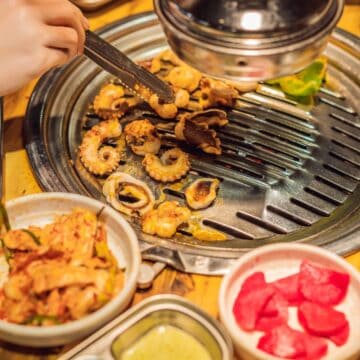 A person grills seafood pieces, possibly octopus, on a tabletop grill surrounded by traditional Korean side dishes like kimchi, pickled radishes, and assorted sauces. The scene is set for a flavorful meal at a Korean barbecue restaurant.