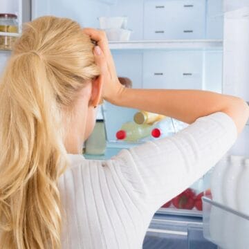 A woman with long blonde hair stands in front of an open refrigerator, holding her head with both hands. Inside the fridge are bottles of milk, a salad bowl, and various jars. She appears to be pondering or frustrated.