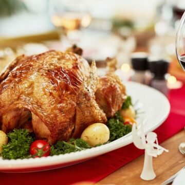 A roasted turkey, one of the unique holiday dinner staples in different countries, sits on a white platter garnished with parsley and small potatoes. Nearby, a glass of red wine graces a wooden table set with a red table runner and various dishes.