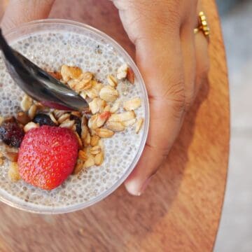 A hand holds a plastic cup of chia seed pudding, rich in fiber foods, topped with granola, a strawberry, and a black spoon. The cup rests on a round wooden surface.
