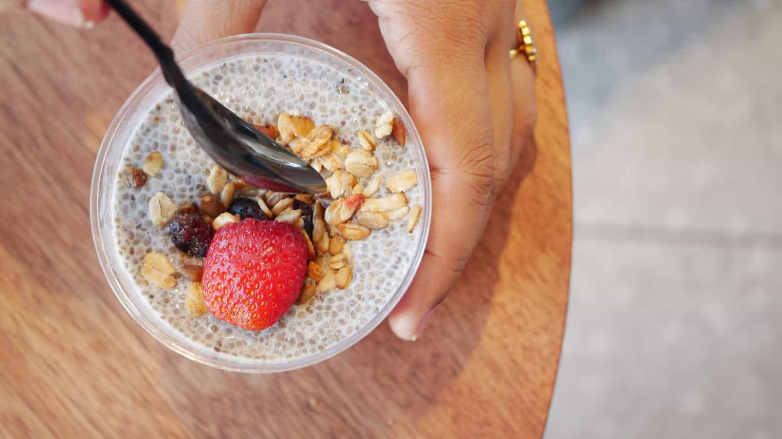A hand holds a plastic cup of chia seed pudding, rich in fiber foods, topped with granola, a strawberry, and a black spoon. The cup rests on a round wooden surface.