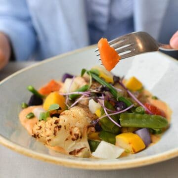 A close-up of a plate filled with a colorful assortment of vegetables, including roasted cauliflower, yellow squash, green beans, and carrots, garnished with fresh microgreens. A hand is holding a fork with a piece of carrot, ready to take a bite. The dish is served on a light, oval plate with a delicate sauce, and the background shows a person in a light blue jacket, blurred to keep the focus on the food.
