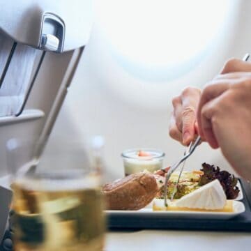 A person is eating a meal on an airplane. The tray table holds a variety of foods, including a salad, bread, and a dessert. A drink in a plastic cup is visible in the foreground. The background shows the airplane window and seat back.