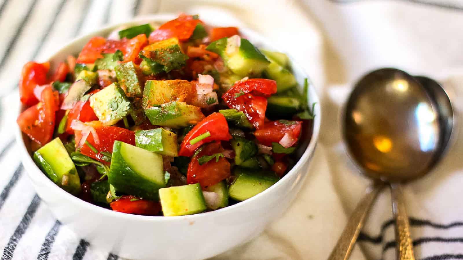 A white bowl brimming with a vibrant salad of chopped cucumbers, tomatoes, and herbs sits elegantly on a striped cloth. Next to it are two metal spoons, poised like an auto draft waiting its turn.