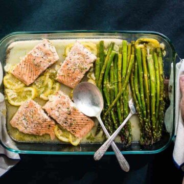A glass baking dish holds four pieces of cooked salmon on lemon slices and a portion of asparagus, perfect for those seeking Thanksgiving seafood dishes. Two hands cradle the dish, and a serving spoon rests on top. The background features a dark surface and a stack of plates with forks.