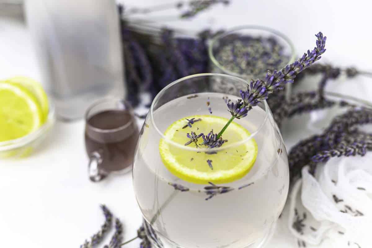 Close-up of a non-alcoholic cocktail garnished with lemon slices and lavender sprigs. Lavender surrounds the glass on the table. A sip of dark liquid and lemon slices in the background create a fragrant and refreshing presentation reminiscent of Grandma's timeless touch.