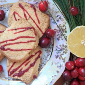 A decorative plate holds four cookies drizzled with red icing, surrounded by fresh cranberries. Beside the plate are lemon halves and additional cranberries. Pine branches are arranged next to the plate.