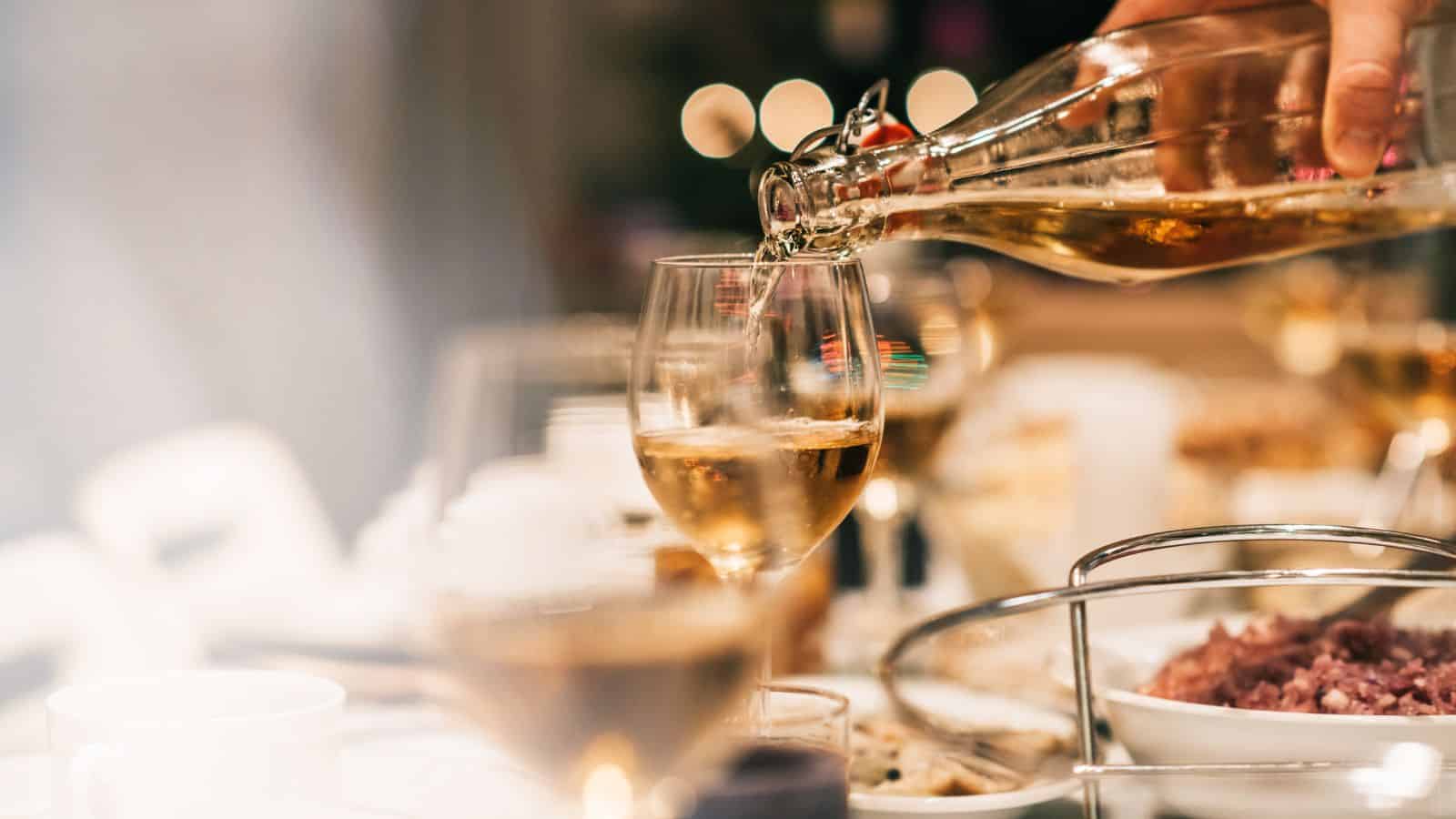 A hand pours champagne into a glass at a dining table set for a festive occasion. Plates, food, and other filled glasses hint at a gathering where healthier holiday feast tips might come in handy for those eager to celebrate with delicious mindfulness.