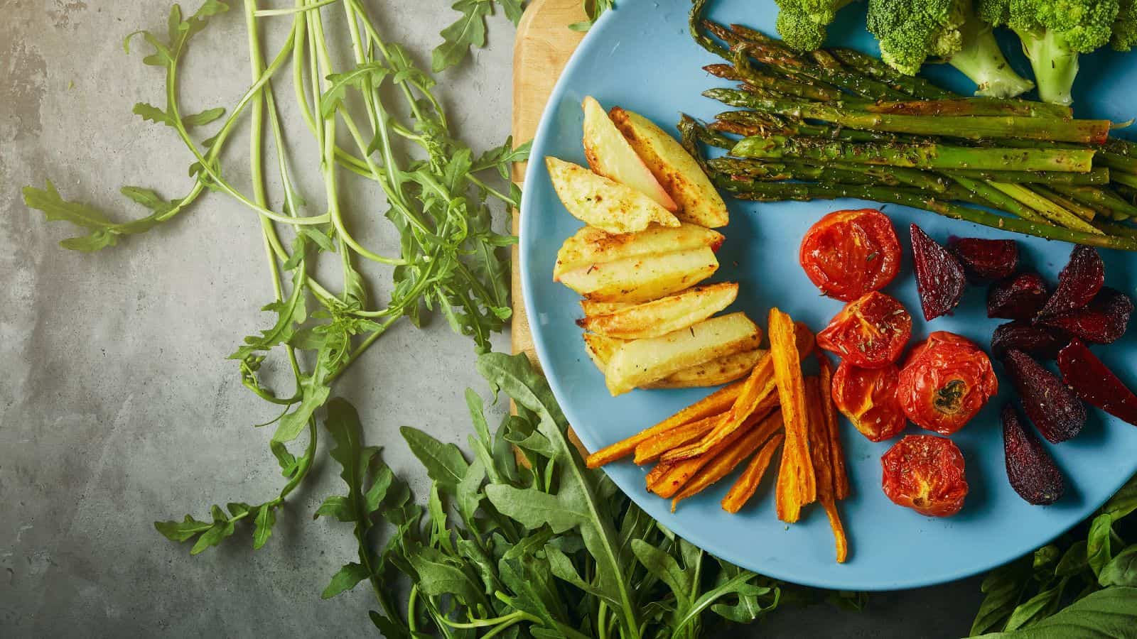 A blue plate with roasted asparagus, cherry tomatoes, beet slices, potato wedges, and carrot sticks on a wooden board, surrounded by arugula leaves. These vibrant veggies are arranged neatly on the right side of the plate—a simple tip for crafting a healthier holiday feast.