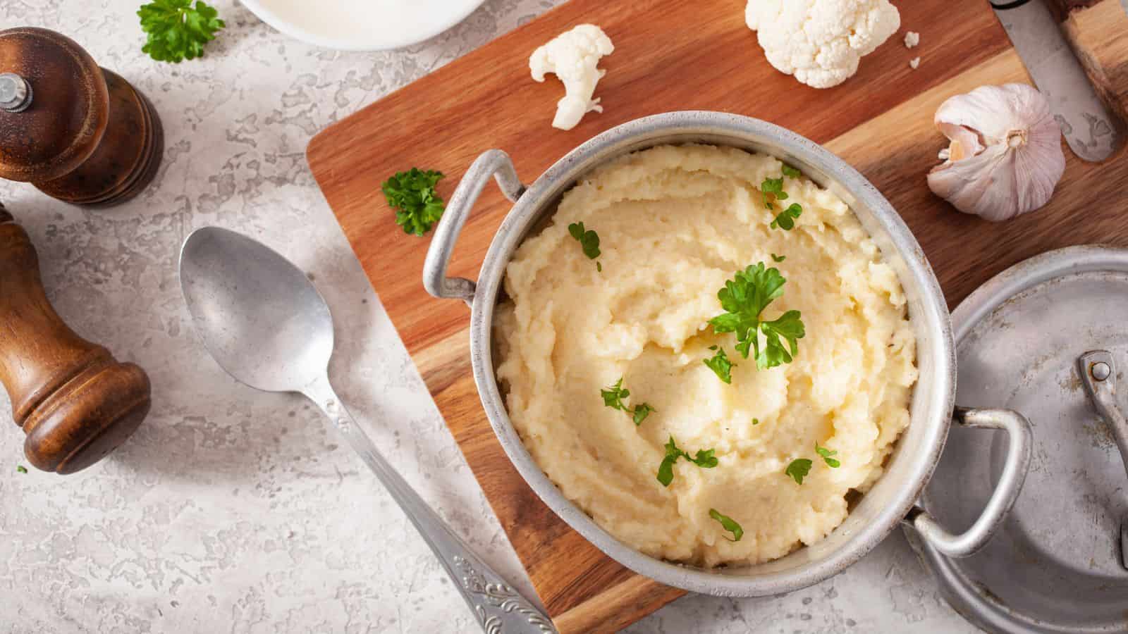 A bowl of creamy mashed cauliflower garnished with parsley sits on a wooden cutting board, perfect for a healthier holiday feast. Nearby are a spoon, garlic, cauliflower pieces, and a black pepper grinder on a textured surface.