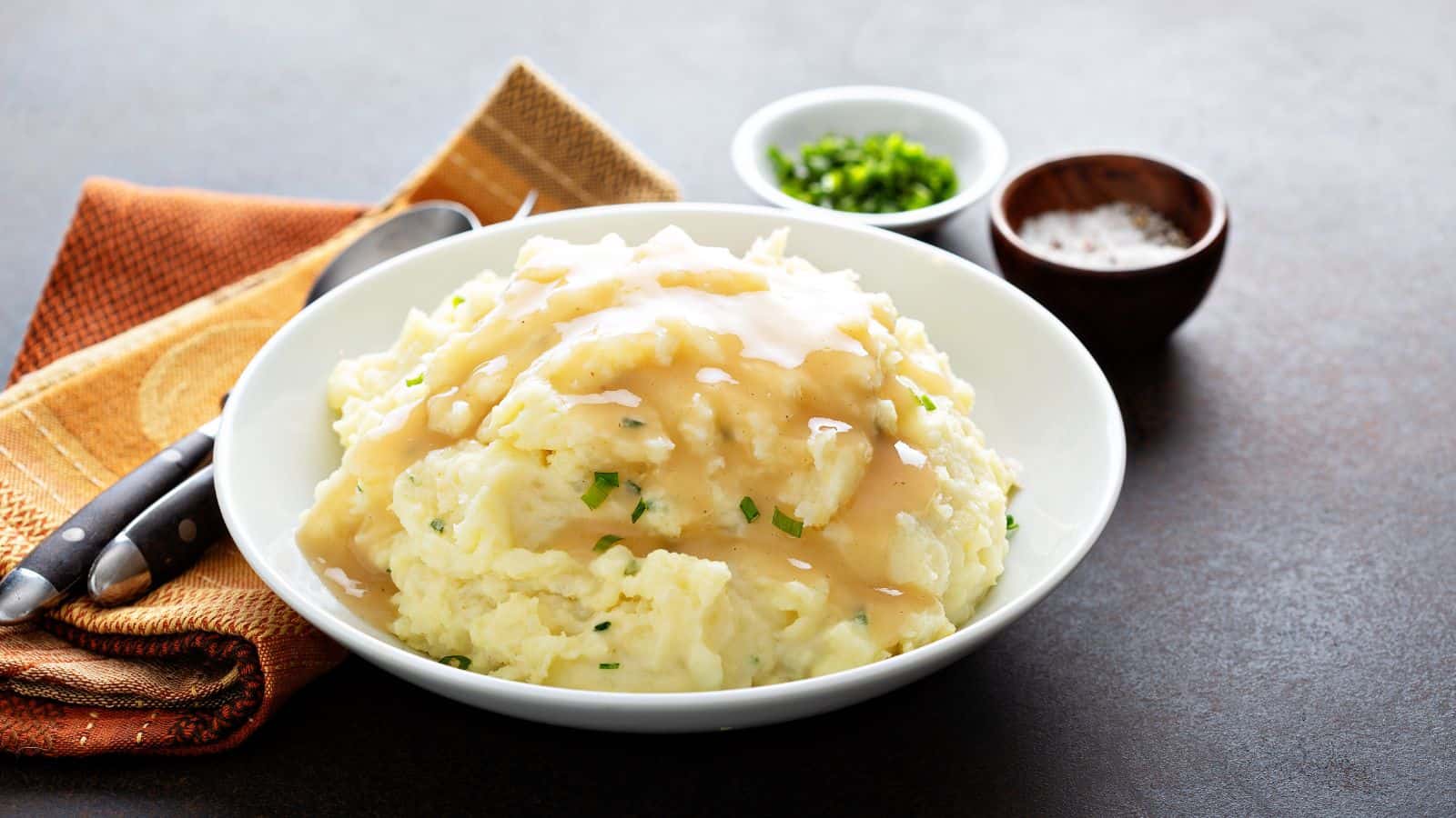 A bowl of mashed potatoes topped with gravy is placed on a table. Nearby, there are small bowls containing chopped green onions and salt. A knife and fork rest on an orange cloth next to the bowl.