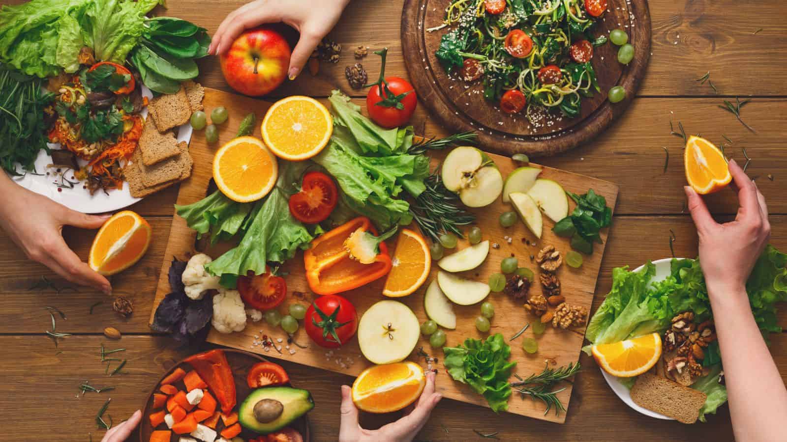 A wooden table covered with fresh fruits and vegetables, including oranges, apples, tomatoes, and leafy greens. Plates with salads and mixed vegetables are present. Three hands are reaching for the food, suggesting a shared meal setting.