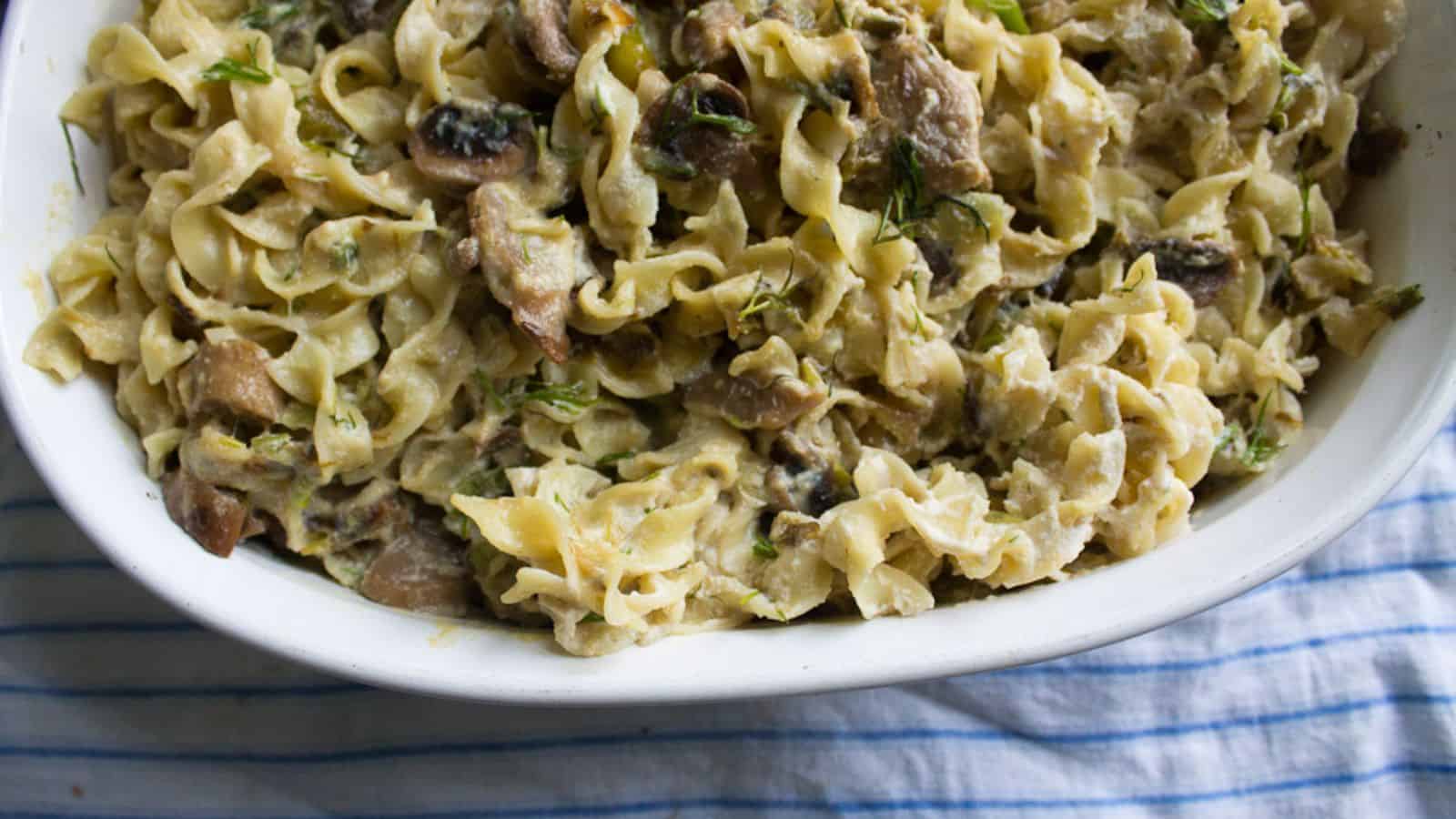 A white dish filled with creamy pasta topped with mushrooms and herbs is placed on a blue and white striped cloth.