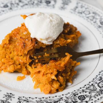 A piece of carrot cake topped with cream is on a decorative black and white plate. A fork is partially inserted into the cake, and part of a mug is visible in the background.