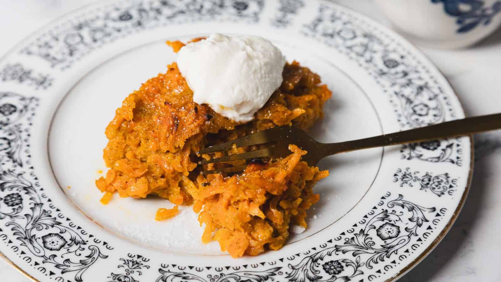 A piece of carrot cake topped with cream is on a decorative black and white plate. A fork is partially inserted into the cake, and part of a mug is visible in the background.