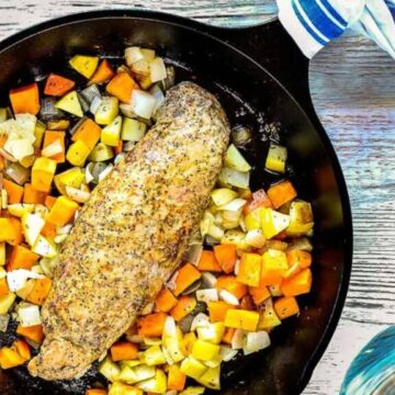 A roasted pork tenderloin rests in a cast iron skillet surrounded by diced, cooked vegetables, including potatoes, sweet potatoes, and onions. The skillet is on a wooden surface near a striped napkin and a blue glass.