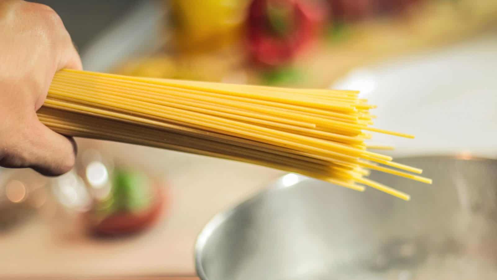 A hand holding uncooked spaghetti over a pot of boiling water, preparing to cook them. The background shows blurred kitchen items, including a red pepper and a yellow surface.