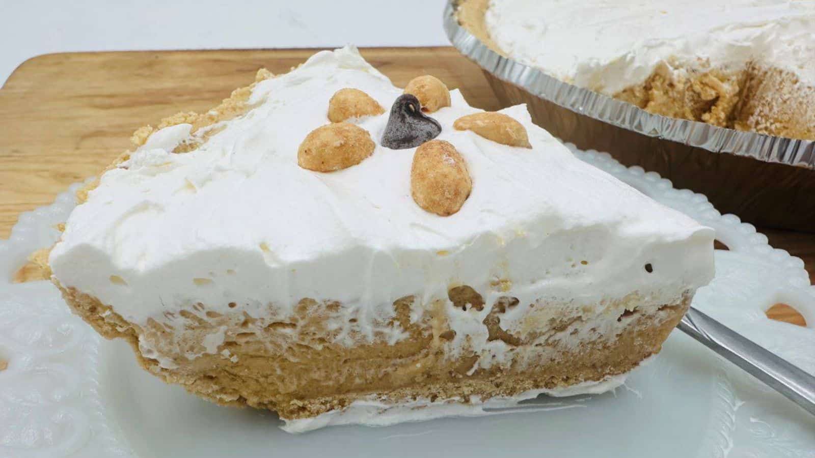 A slice of peanut butter pie topped with whipped cream and peanuts, on a white plate with the whole pie in the background.