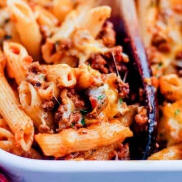 A close-up of a baked pasta dish with penne, ground meat, and melted cheese in a white rectangular dish. A wooden spoon is partially visible, serving the pasta. The background features a red and white checkered cloth.