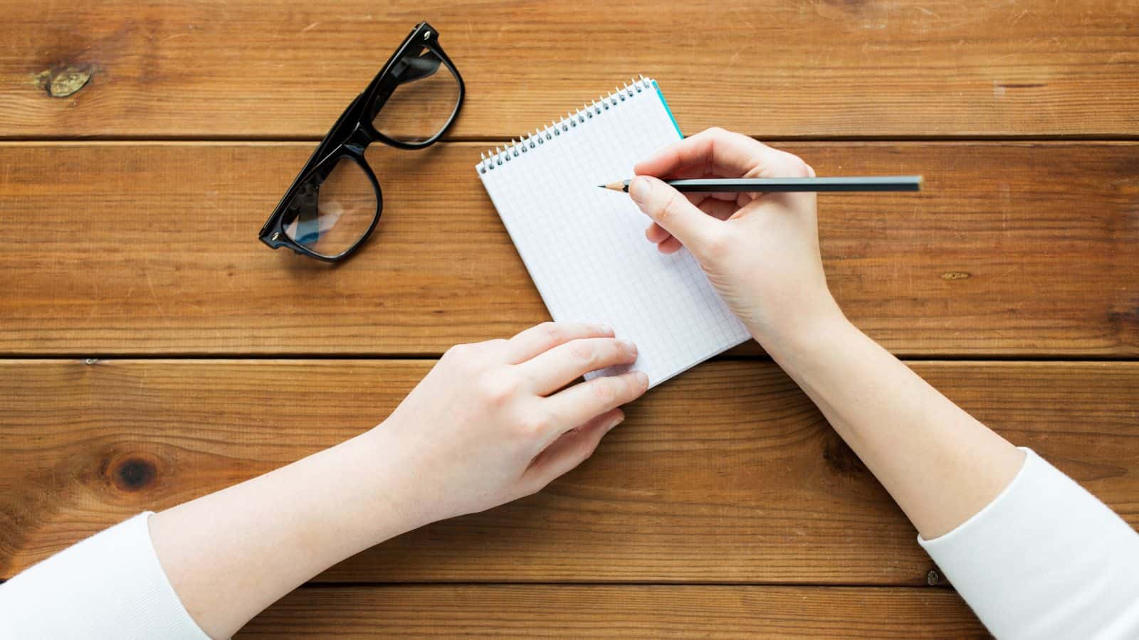 A person writes on a small spiral-bound notepad with a pencil on a wooden table, jotting down holiday meal planning tips. A pair of black-rimmed glasses is placed nearby. The person, dressed in a white long-sleeve shirt, appears deep in thought as they organize their festive feast ideas.