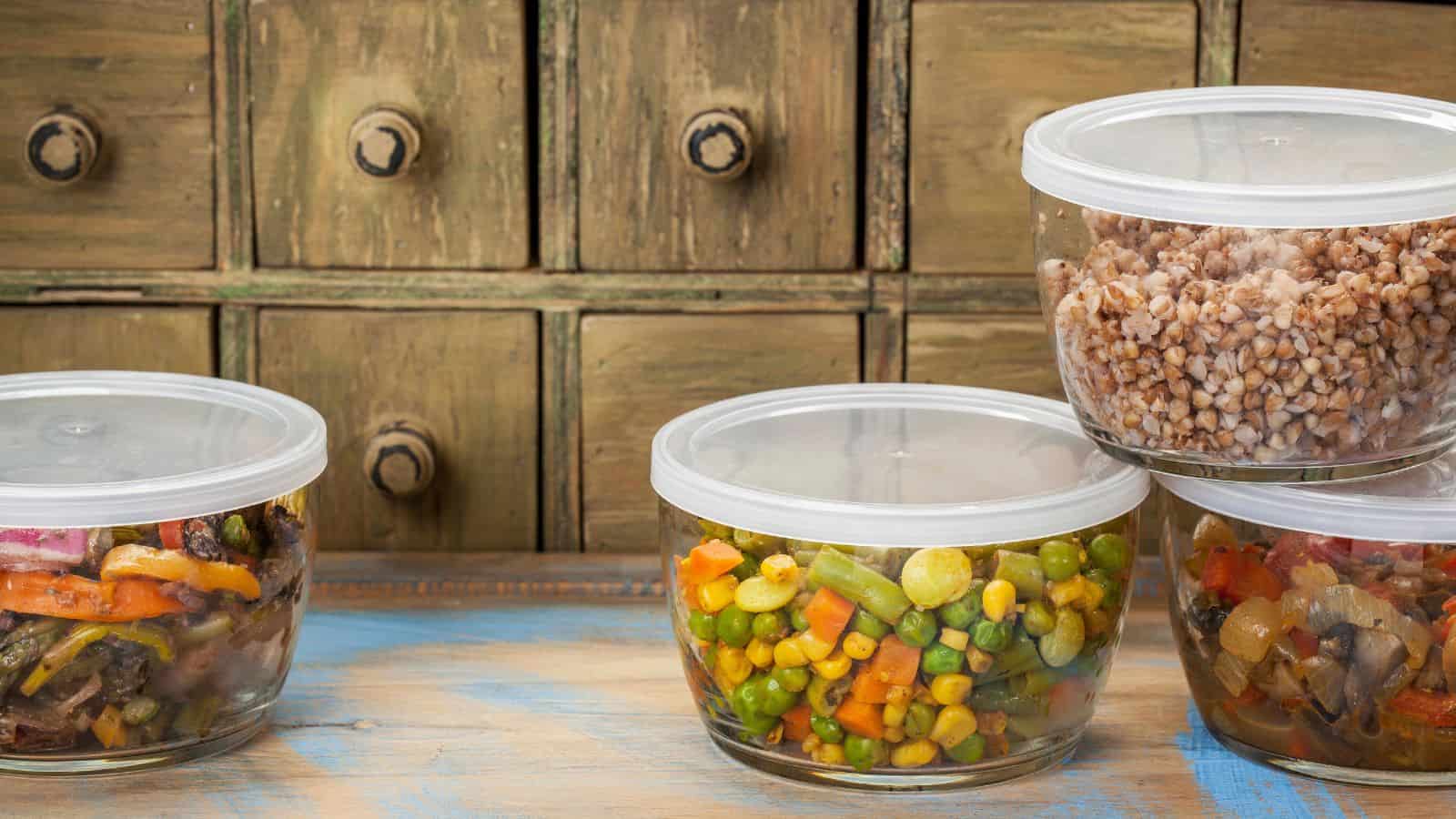 Four transparent containers with lids, filled with different types of food like mixed vegetables and grains, sit on a wooden surface. With a rustic wooden cabinet as the backdrop, the scene offers simple tips for creating a healthier holiday feast that delights both the eyes and palate.