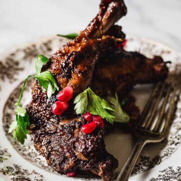A plate of grilled chicken garnished with fresh parsley and red pomegranate seeds. The chicken is served on a decorative floral-patterned plate with a fork on the side. The background is a marble surface.