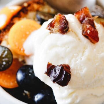 A dessert bowl with a scoop of vanilla ice cream topped with chopped dates. Next to the ice cream are blueberries and halved golden berries. A spoon is placed in the bowl, which is set on a blue textured surface.