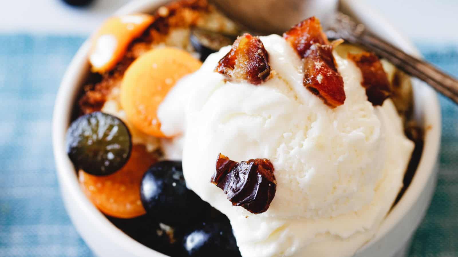 A bowl of dessert features a scoop of vanilla ice cream topped with pieces of chopped dates. Surrounding the ice cream are slices of yellow and purple fruits, along with dark grapes. A spoon rests in the bowl.