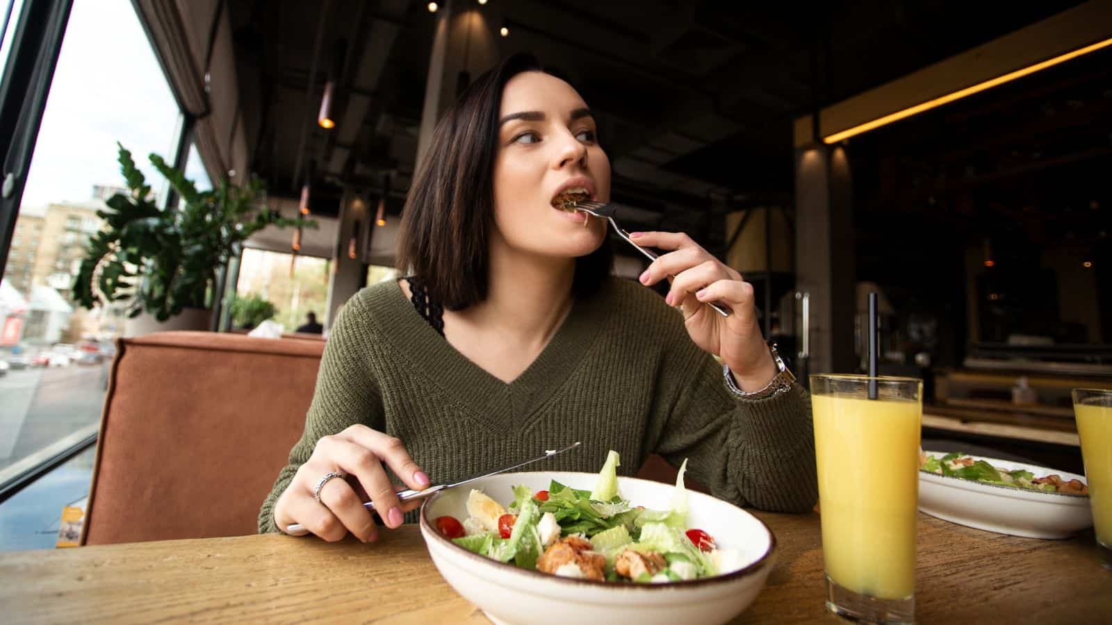 A woman in a green sweater enjoys a salad at a wooden table in the restaurant, savoring her healthier holiday feast. She takes a sip of orange juice, surrounded by large windows and lush plants, creating an airy ambiance.