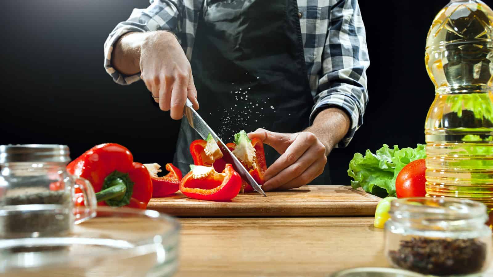 A person in a checkered shirt and apron is slicing a red bell pepper on a wooden cutting board. As they prepare, surrounded by kitchen items—a lettuce head, tomato, glass bowl, and bottle of oil—they reflect on holiday meal planning tips for festive feasts.