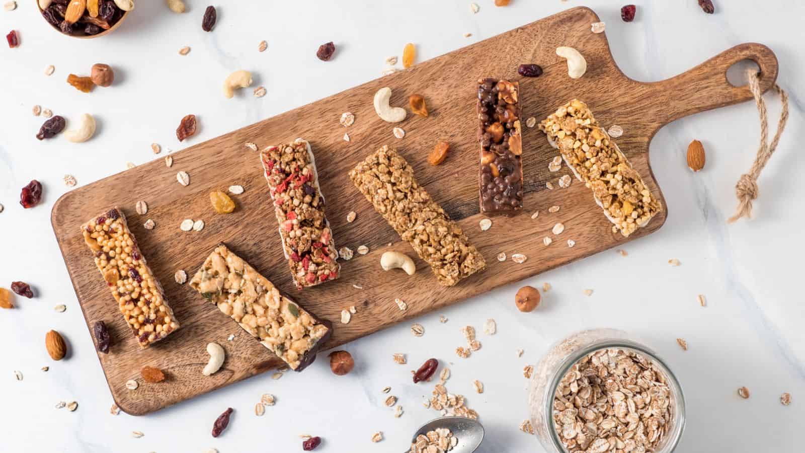 A wooden board displays six granola bars with various ingredients, surrounded by scattered nuts, raisins, and oats on a light surface. A jar filled with oats and a spoon are placed nearby.