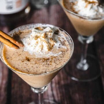 Two martini glasses filled with a creamy pumpkin spice beverage topped with whipped cream and sprinkled with cinnamon. A cinnamon stick rests on the rim of each glass. A blurred background includes spices and a cookie.