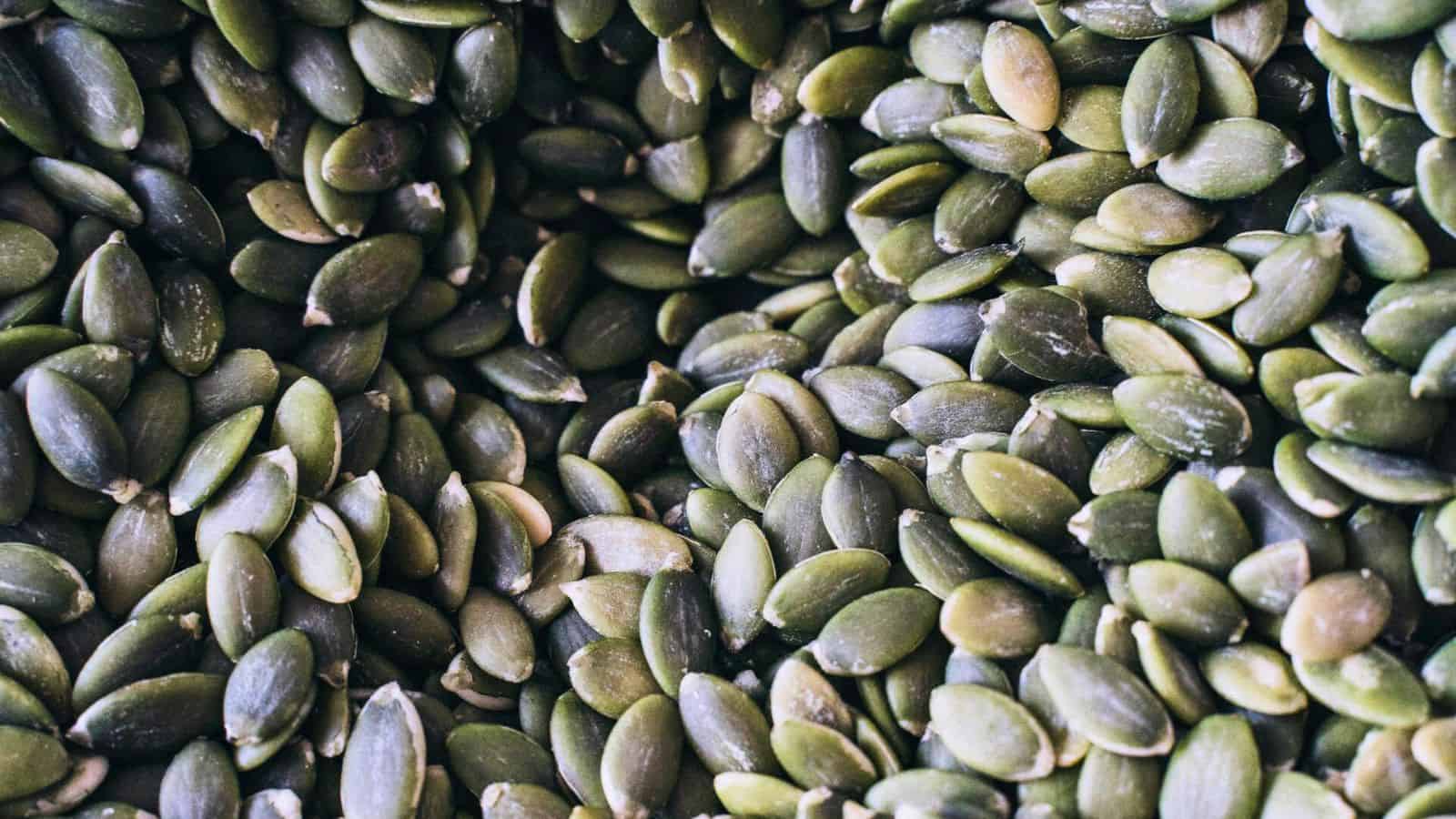 A close-up view of a pile of green pumpkin seeds with visible texture and details. The seeds are layered closely together, showcasing their elongated, oval shapes and smooth surfaces.