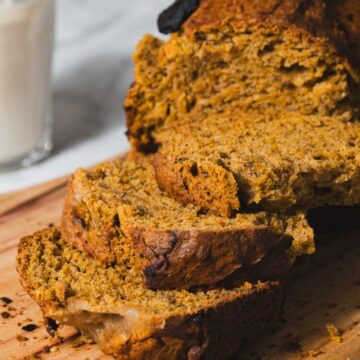 Slices of a brown loaf of bread sit on a wooden cutting board. A glass of milk is in the background.