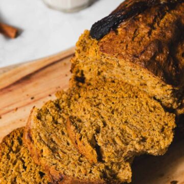 A loaf of homemade pumpkin bread is partially sliced on a wooden cutting board. The bread has a golden-brown crust, and two cinnamon sticks are visible in the background.