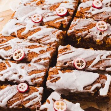 Sliced dessert bars topped with white icing and small red and white decorative slices are arranged on a wooden cutting board. The bars have a brown, spiced appearance, hinting at flavors like gingerbread or pumpkin spice.