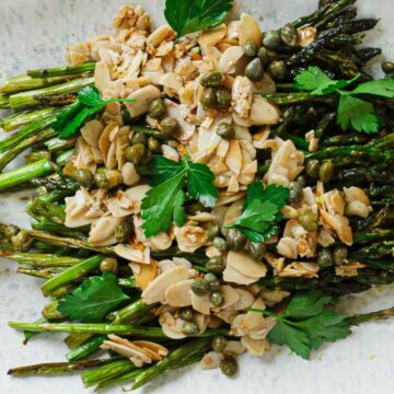 A plate of roasted asparagus topped with sliced almonds, capers, and fresh parsley. Two lemon wedges are placed on the side for garnish.