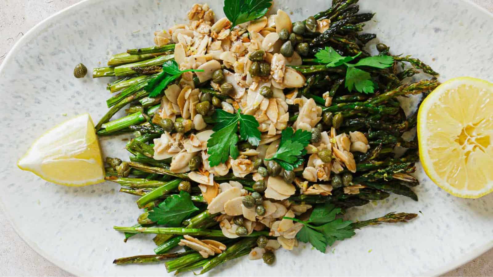 A plate of roasted asparagus topped with almond slices, capers, and parsley. Two lemon wedges are placed on either side of the asparagus. The dish is presented on an oval white plate.