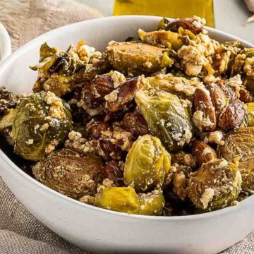 A bowl of roasted Brussels sprouts with almonds, crumbled cheese, and seasoning sits on a cloth. A small bowl of dressing and a dish with herbs are nearby on a wooden surface.