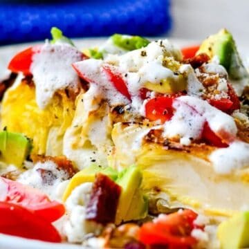 A plate with a colorful dish featuring a large wedge of iceberg lettuce topped with diced tomatoes, avocado pieces, crumbled cheese, bacon bits, and drizzled with creamy dressing. A blue napkin is in the background.