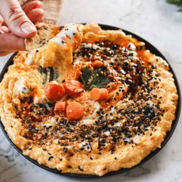 A person holds a piece of bread topped with dip above a bowl of creamy orange hummus garnished with carrots, seeds, and herbs on a marble surface. A small dish of seasoning and fresh green herbs are nearby.